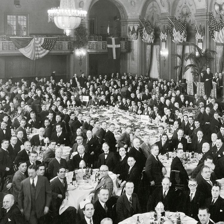 Black and white image of people in formal business attire in a grand banquet hall.