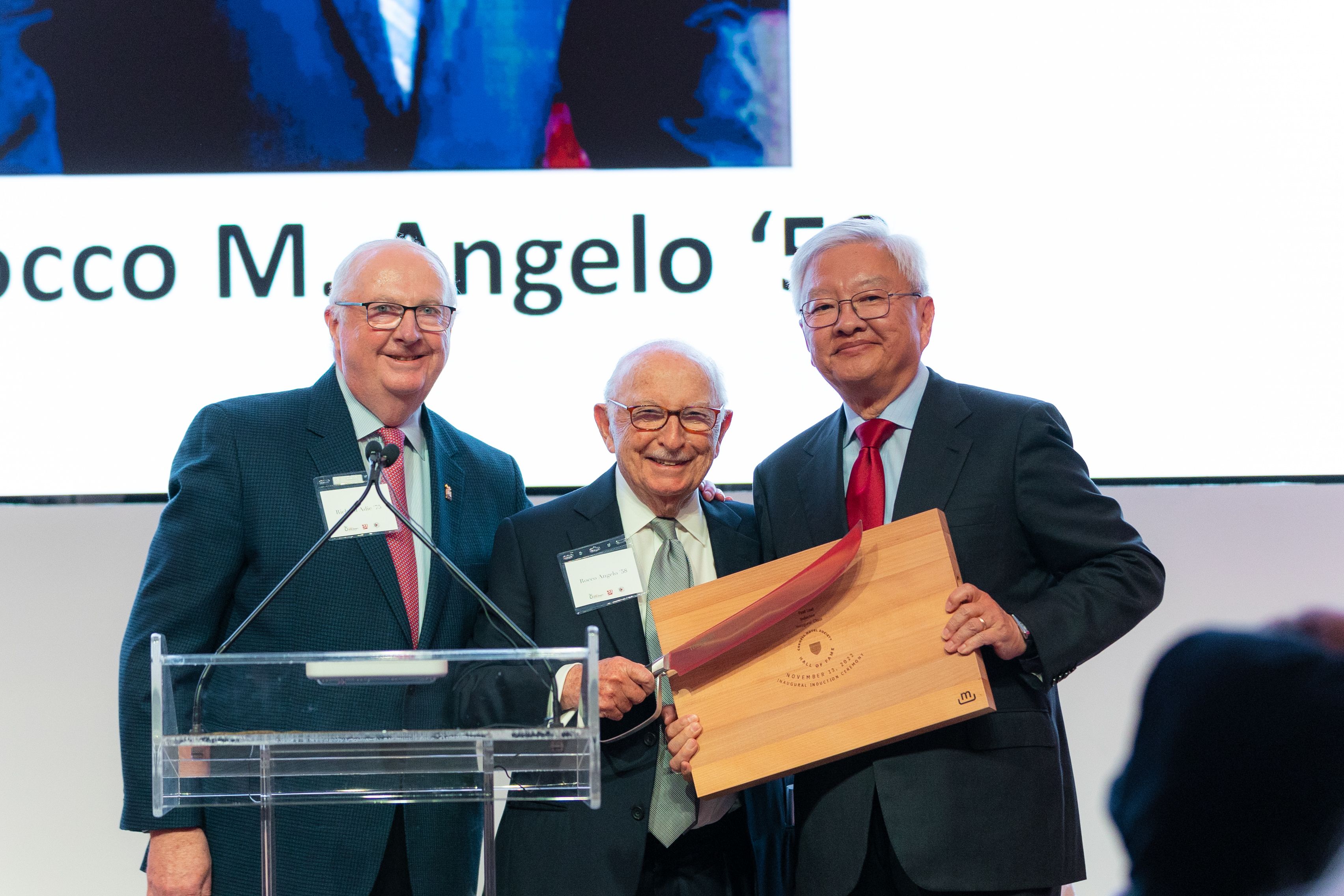 Three men in suits pose together behind a clear podium holding a wooden cutting board and knife. 