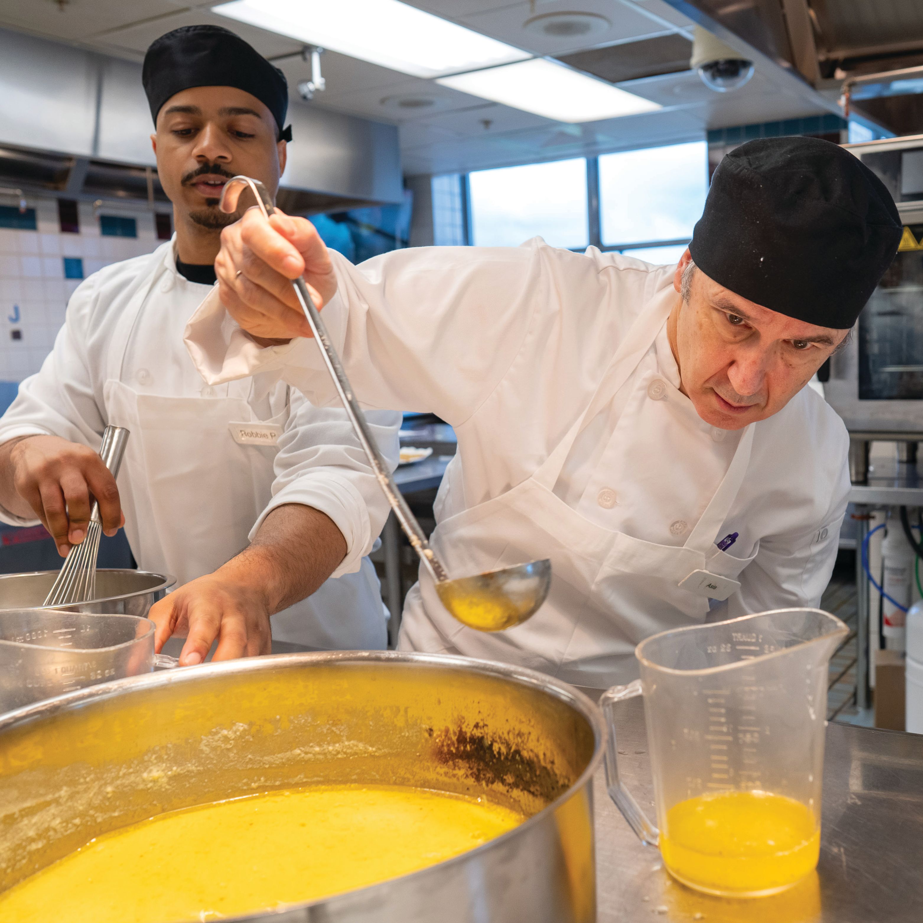 Asís Martínez-Jerez wears a chef uniform and holds a ladle next to a large pot of yellow hollandaise.