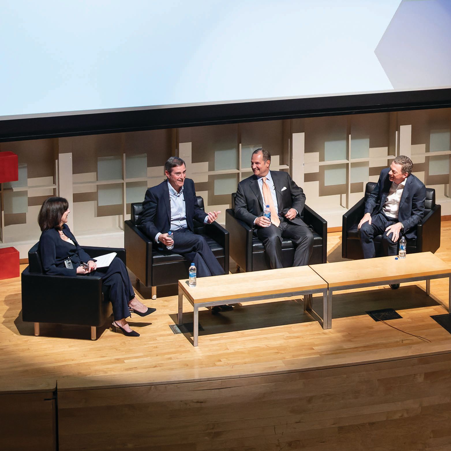 Four people sit in armchairs on a stage in front of a projector screen.