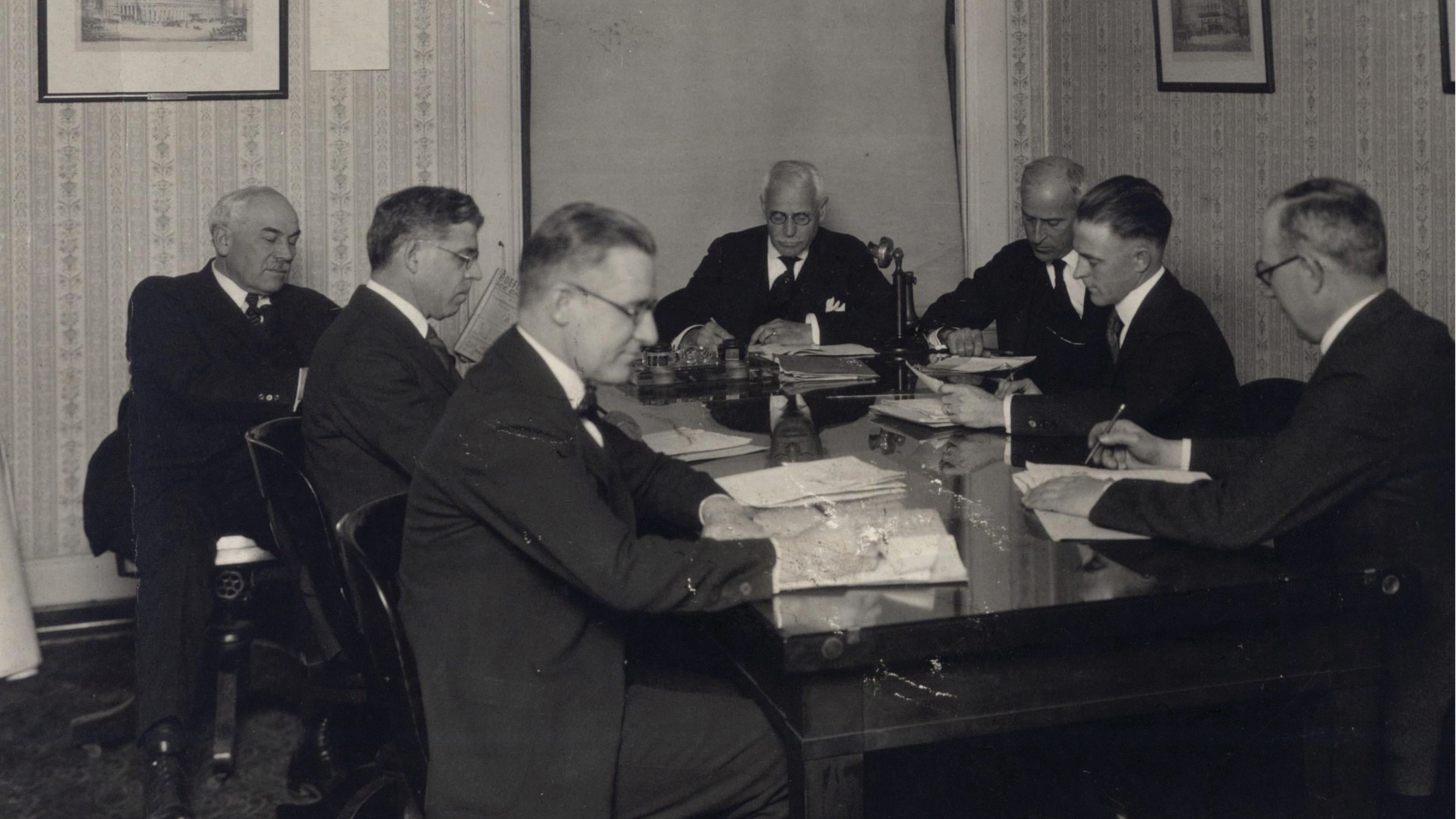 Men in business suits sit at a conference table.