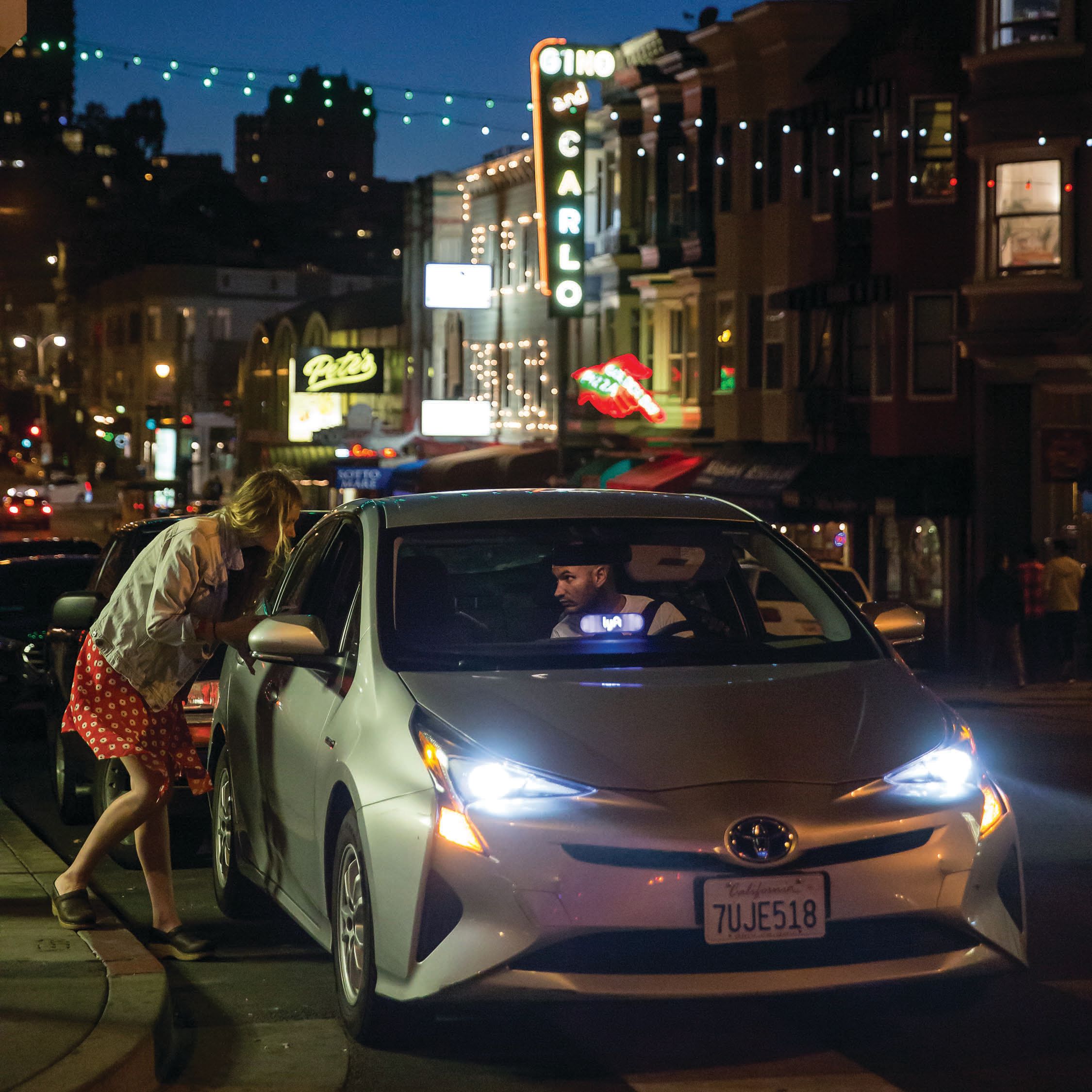A woman leans into the passenger side window of a car to speak to a driver.