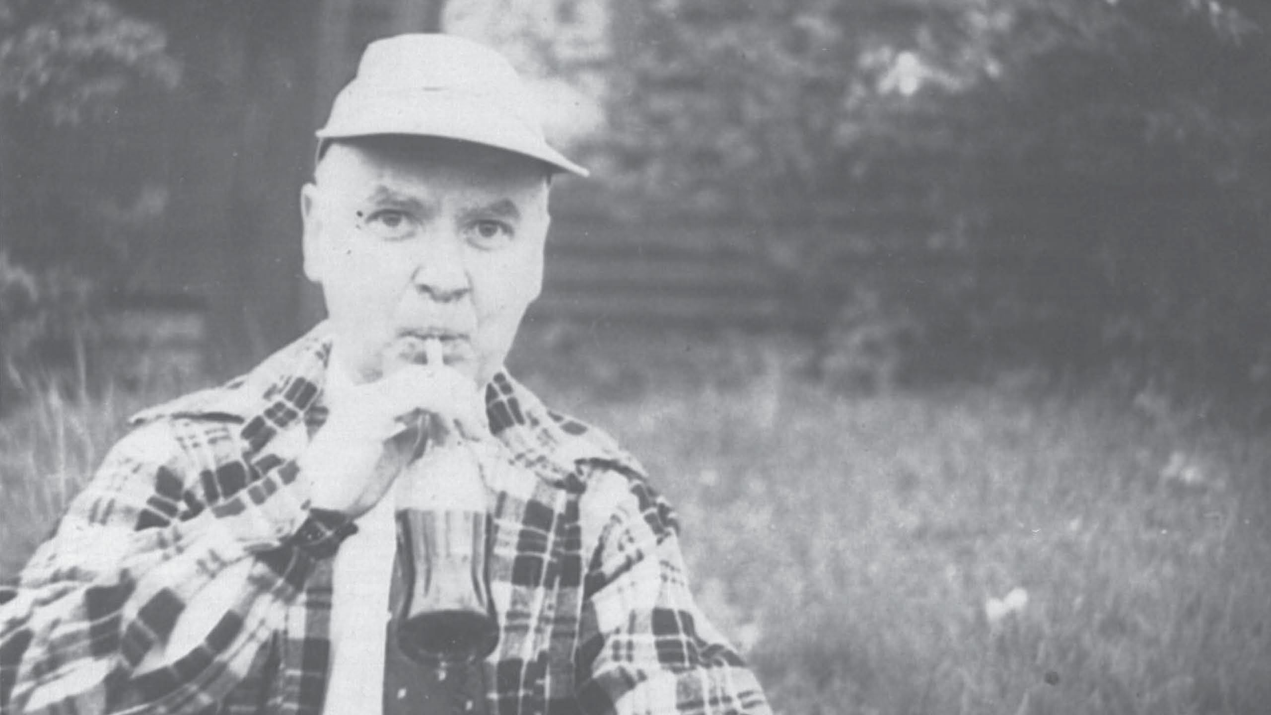 Dean Meek sips a glass bottle of soda in a flannel shirt and baseball hat.