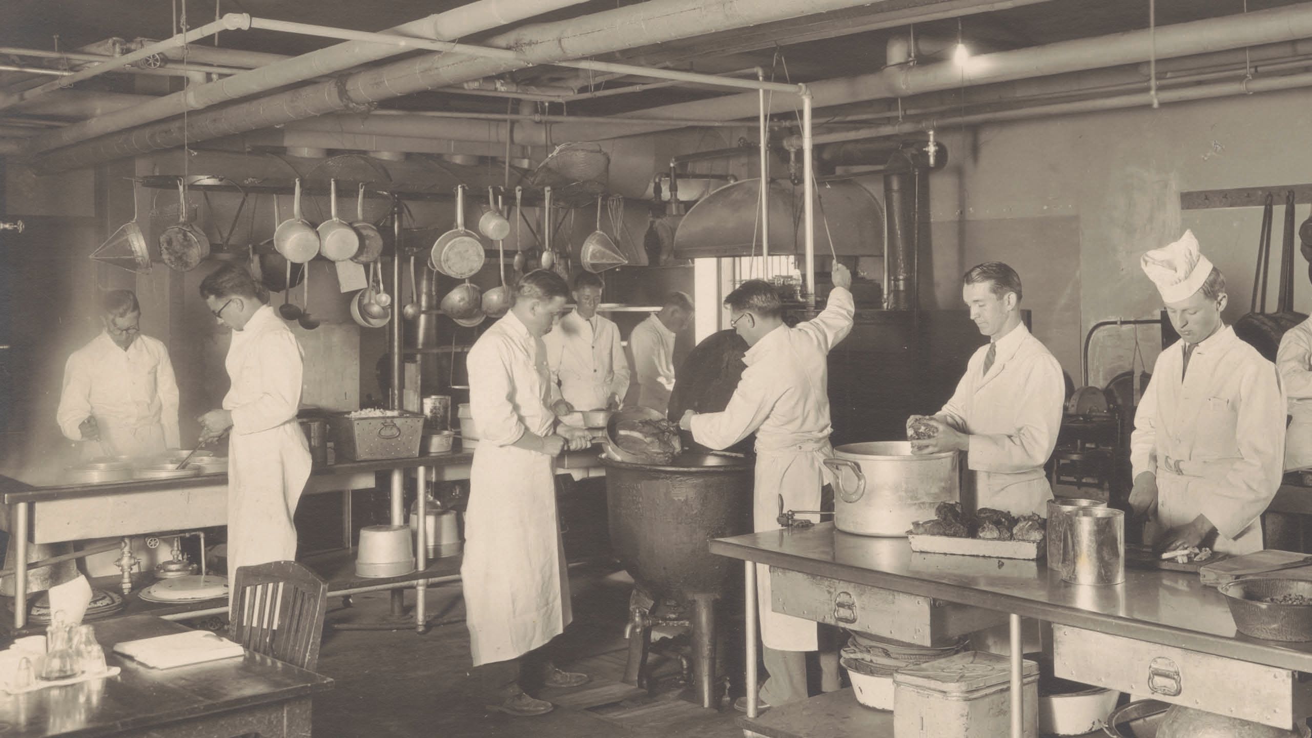 Black and white photo of students in chef clothing working in a kitchen.