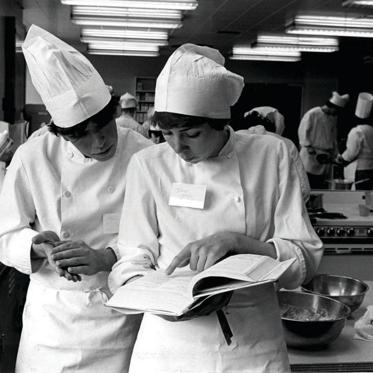 Two students in chef uniforms review a cookbook.