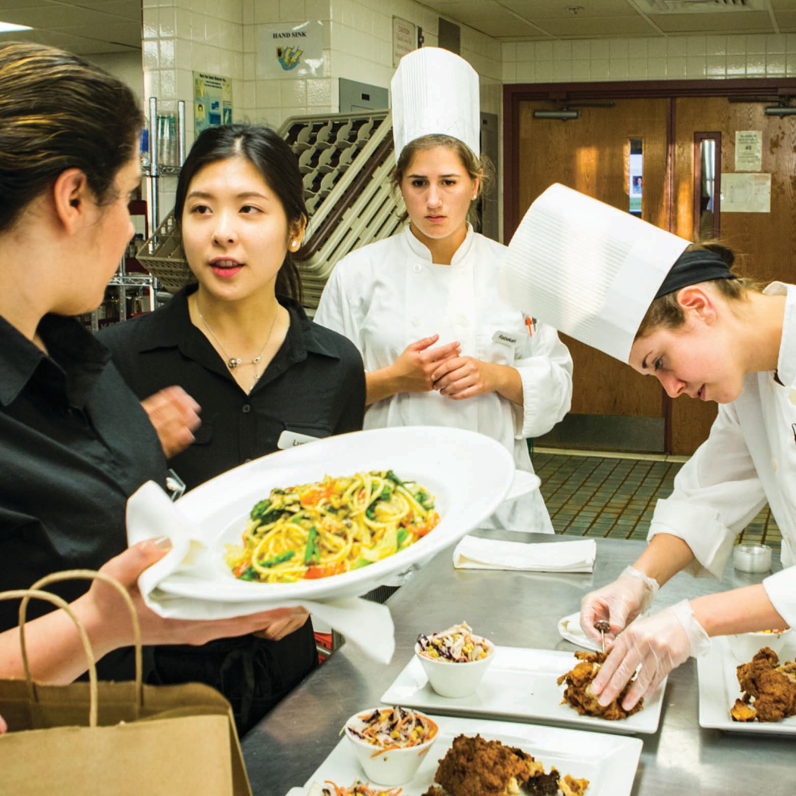 Student chefs and servers in the kitchen prepare and grab dishes.